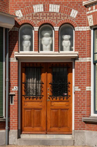 A beautifully varnished double wooden door in a chic, old building (probably around 1920). On both sides are walls with small, orange-brown stones, interspersed with rows of white stones. Above the door, three narrow, arched windows stand side by side. Behind each window is a large, white plastic bear. The largest is in the center, and two smaller ones are on the left and right. They are almost as large as the window.