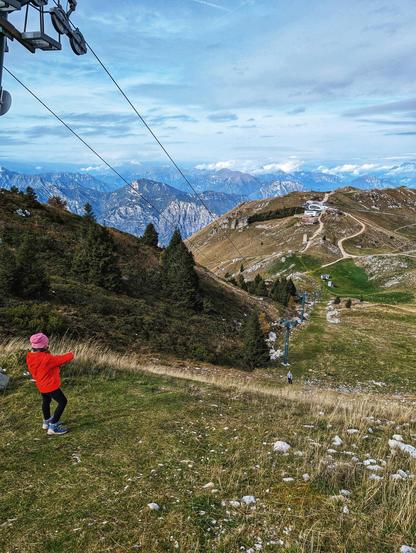 A high-angle shot of a young person in a red jacket and pink hat, viewed from behind, standing on a grassy mountain slope. They are pointing towards a stunning panoramic view of the mountains. A ski lift cable runs across the top of the image and down the slope. In the mid-ground, a second person is visible at the bottom of the slope, wearing a grey jacket. In the background, there is a second, smaller chairlift, and a few buildings, namely the summit station of the Funivia Malcesine/Monte Baldo cable car, are visible on the far side of the mountain. The sky is blue with scattered clouds, and the distant mountain ranges have a light dusting of snow.
