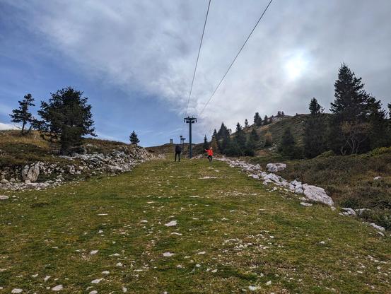 A low-angle, wide shot of a grassy ski slope on a mountain, looking upwards towards the summit. Two figures are walking up the slope; one in dark clothing and another in a bright red jacket. A ski lift cable stretches overhead, supported by a large pylon in the middle distance. The slope is lined with scattered rocks and evergreen trees on either side. The sky is partly cloudy, with the sun partially obscured by clouds.