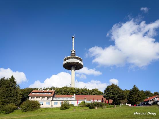 Der 144 Meter hohe Funkturm auf dem Hoherodskopf ragt über Bäume und Gasthäuser hinaus in den klaren blauen Himmel. Ein Wahrzeichen, das Technik und Natur in der Vogelsberglandschaft verbindet.