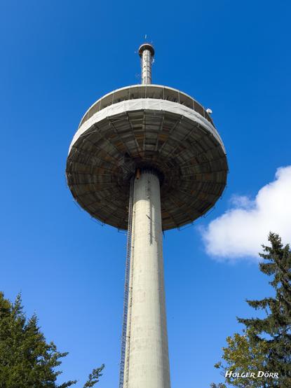 Blick aus der Froschperspektive auf den Funkturm Hoherodskopf: Die gewaltige Betonröhre trägt die runde Plattform und die Antenne, die sich kontrastreich gegen das tiefe Blau des Himmels erhebt – monumental und futuristisch zugleich.
