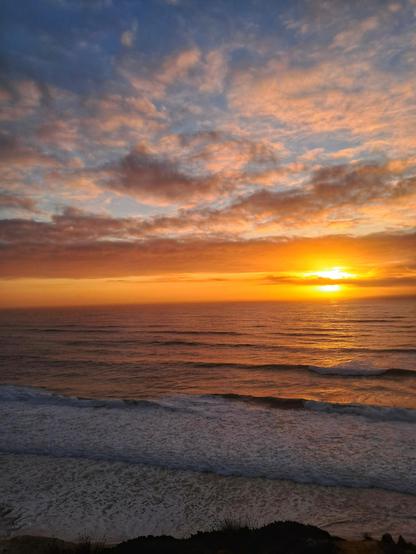 [pt] Pôr do sol sobre o mar com ondas em primeiro plano. O sol laranja brilha atrás de nuvens com o céu azul em fundo

[en] Sunset over the sea with waves in the foreground. The orange sun shines behind clouds with the blue sky in the background