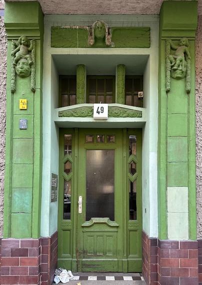 An ornate green doorway with Jugendstil and Art Deco elements. The number above the door reads 49. The walls framing the doorway are decorated with reliefs of cherubic figures.