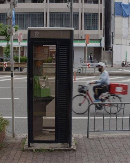 Empty telephone both by the side of a road, with a man passing in the background.