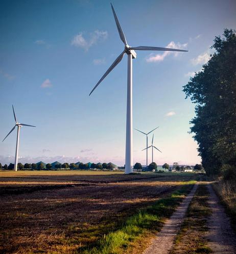 Windkraftwerke auf Ackerflächen. Die Rotoren zeigen dir entgegen, also hast du Rückenwind beim Fahrradfahren.
Am Horizont sieht man eine Biogas Produktionsanlage.