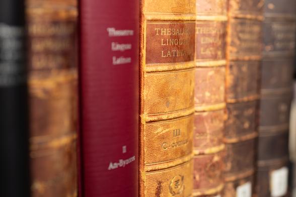 The image shows a row of old, hardbound books on a shelf. The book spines are of various colours, including shades of brown, red, and black. The focus is on a reddish-brown volume with the title “Thesaurus Linguae Latinae” in gold lettering. The other books, with faded lettering and worn textures, are slightly out of focus, suggesting age and frequent use. The composition captures a sense of history and scholarly inquiry.