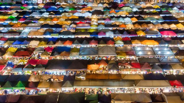 An aerial view of a vibrant night market with numerous colorful stalls and tents. The stalls are illuminated, showcasing a variety of goods and a lively crowd of people.