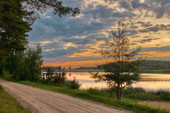 A serene landscape featuring a dirt road alongside a calm lake, bordered by lush greenery and trees. The sky is painted with soft hues of orange and yellow as the sun sets behind clouds, creating a tranquil atmosphere.