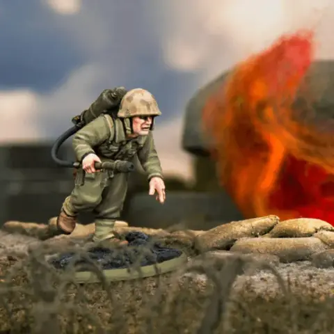 A detailed miniature of a U.S. soldier, Hershel W. 'Woody' Williams, is depicted in action, equipped with a flamethrower on his back and wearing a helmet and military uniform. He is positioned on a textured base that resembles a battlefield, with a backdrop of flames and smoke suggesting a combat environment. The scene captures the intensity of the Pacific theatre during World War II.