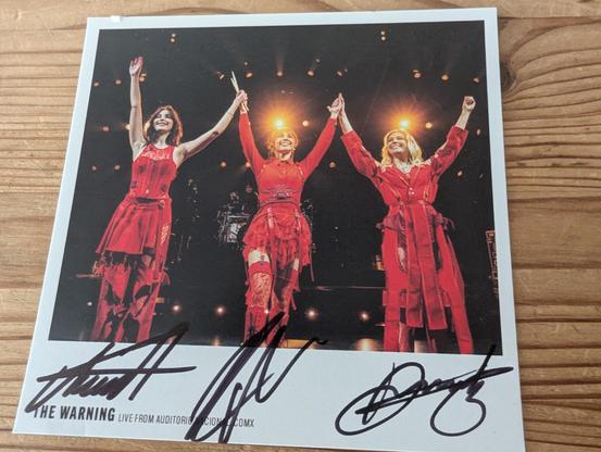 Paper card on a wooden table, showing the band members, on stage, dressed in red, greeting the audience (behind the camera).
Below, signatures from all three