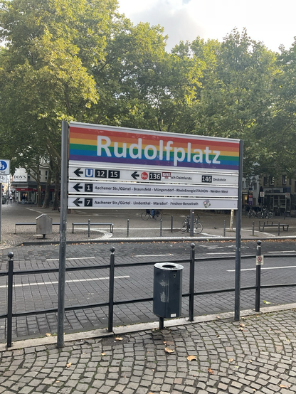 Verkehrsschild „Rudolfplatz“ der KVB in Regenbogenfarben. Dahinter eine leere Straße und einige Bäume