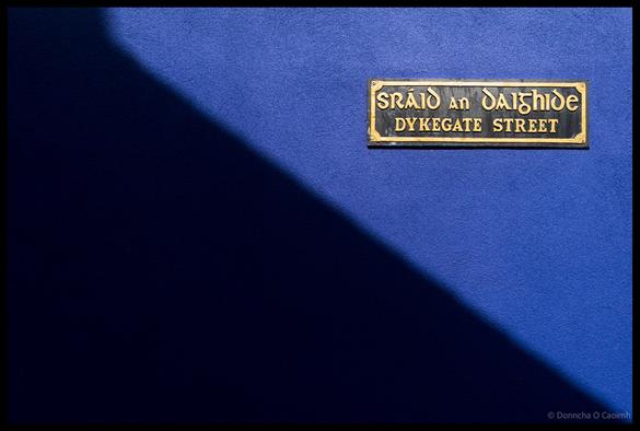 Bilingual street sign reading "SRÁID AN DAIGHIDE / DYKEGATE STREET" mounted on a vibrant blue textured wall with dramatic shadow cast across the surface in Dingle, County Kerry