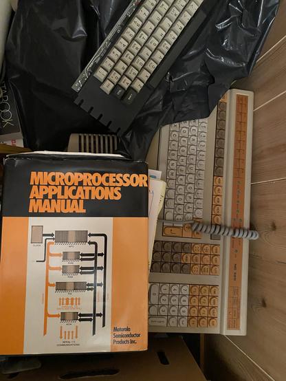 A big orange colored bound bool rests on top of something. Beside it a large beige keyboard with orange, grey and beige keys can be seen resting against a wall. It has Swedish keyboard layout and extra shortcut keys for a word processor. At the top part of the image a smaller computer keyboard  without housing is partially seen.