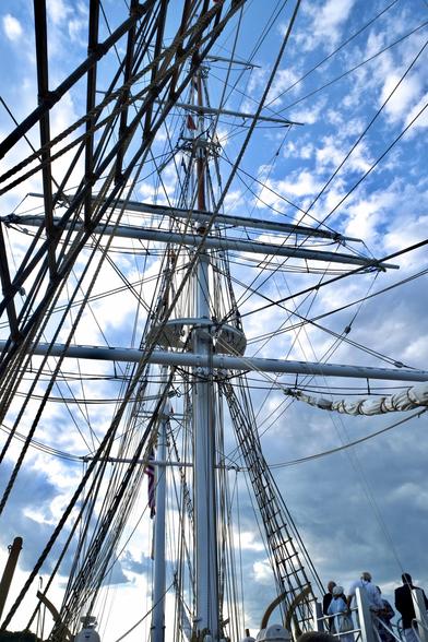 Color photo — deck to top of mainmast — of a wooden whaling ship built in 1841. The bright blue August sky has small scattered clouds, seen from the bow of the ship, through the rigging of the forward mast. All sails are furled. A small group of visitors are about to step onto the deck from a boarding platform at the lower right. The Charles W. Morgan is maintained as a seaworthy vessel, last sailed on its 38th voyage in 2014, Mystic Seaport Museum, Mystic CT USA.