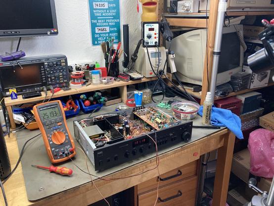 A NAD 7020e receiver with the case removed on my workbench. Several tools and a pile of old capacitors are scattered across the desk. My multimeter is connected to some test points on the circuit board and showing the idling current.