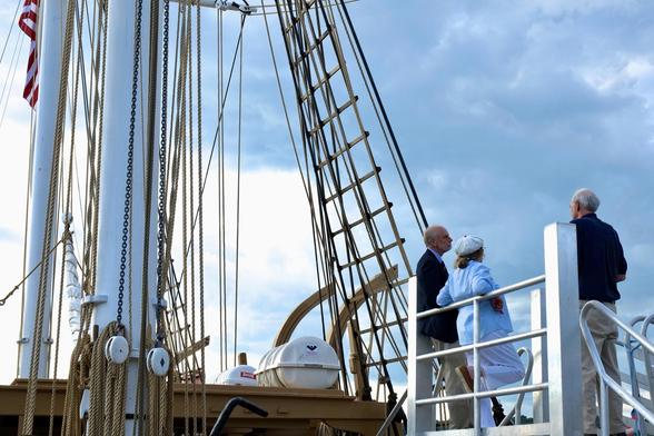 Color photo. Three well dressed visitors relax while standing on a boarding platform at deck level of the Charles W. Morgan — an 1841 wooden whaling ship. The ship is docked at Mystic Seaport Museum, Mystic CT USA. It is late afternoon on a partly cloudy August day, Half the sky is bright blue, but staring to fade.