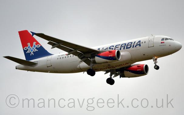 Low side view of a twin engined jet airliner flying from left to right at a very low altitude, with undercarriage lowered and flaps extended from the rear of the wings.
The plane is mostly white, with blue, billboard-style "Air Serbia" titles on the forward fuselage, partially obscured by the wings, repeated in red under the body.
The black registration "YU-APC" is on the upper rear fuselage next to a red, blue, and white Serbian flag.
The top of the tail is red, with the base being blue, with a white scrollwork two-headed eagle with a pale blue chest overlaid on the 2 colours.
The engine pods under the wings have a red top, white bottom, and diagonal blue tripe in the middle.
Flat grey sky fills the background.