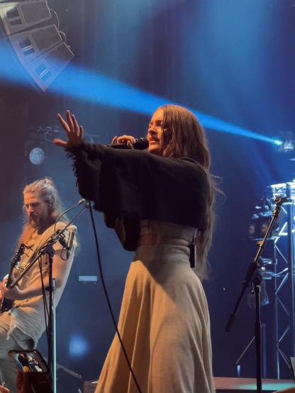 The singer of Kalandra singing on stage and pushing her arm forward in slow motion. In the background the bassist playing his instrument.