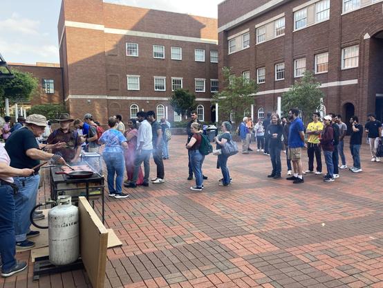 A red brick courtyard with a line of students in front of a large gas grill topped with burgers and hot dogs.