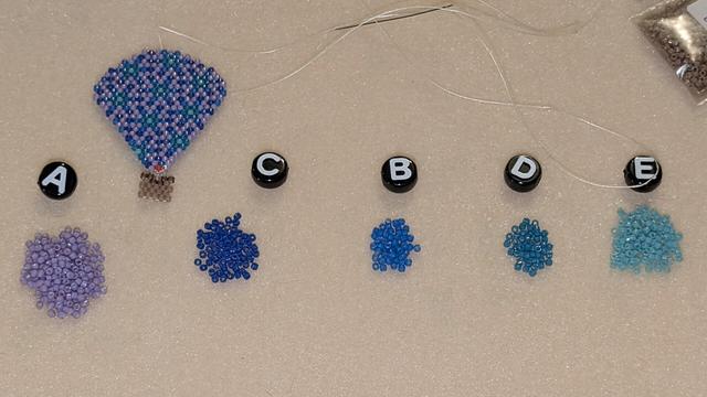 Five colors of delica beads organized on a beading mat; each one has a matching alphabet letter bead to help me match my chosen colors to their placement in the original pattern. There is also a nearly finished balloon earring up in the top left area of the photo. This photo was taken with flash.