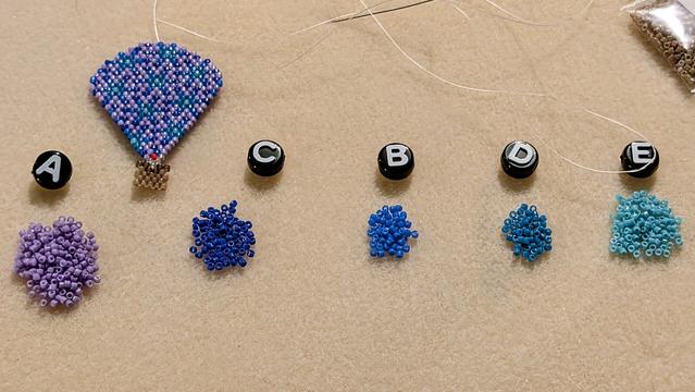 Five colors of delica beads organized on a beading mat; each one has a matching alphabet letter bead to help me match my chosen colors to their placement in the original pattern. There is also a nearly finished balloon earring up in the top left area of the photo. This photo was taken without a flash.