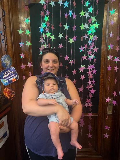 A fat white woman holding a chubby Asian baby in a blue onesie in front of a curtain of purple stars on a wooden door.