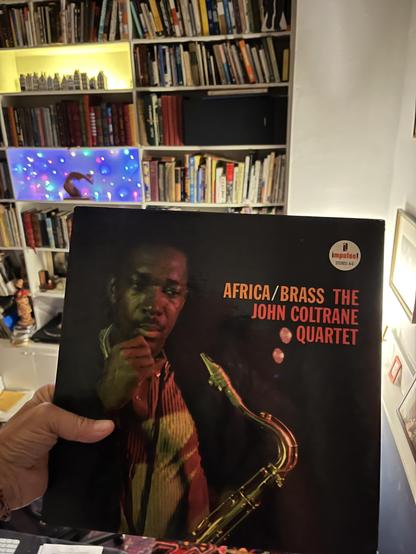 This photo shows someone [it me!] holding up a vinyl record album in what a home library. The album is "Africa/Brass" by The John Coltrane Quartet, released on the Impulse! record label (you can see the distinctive Impulse! logo in the upper right corner). The album cover features a striking image of John Coltrane playing his saxophone, photographed in dramatic lighting that gives his face and instrument a golden glow against a dark background.

Behind the record, you can see extensive bookshelves filled with books, and there are some colorful LED lights creating a festive atmosphere - one shelf has warm yellow lighting, and another has multicolored lights. The setting suggests this is a well-read household with an appreciation for both literature and jazz music. [oh you flatterer you!]

"Africa/Brass" was one of Coltrane's significant albums from 1961, representing his exploration of African musical influences and featuring arrangements by Eric Dolphy. It's considered an important work in Coltrane's discography during his period with Impulse! Records.
