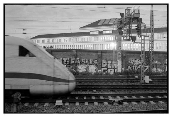 Black and white photograph in landscape orientation. An ICE train is entering the picture in front of a wall covered with graffiti. In the background a large house with a long range of windows. The sun is reflected in some of these windows.