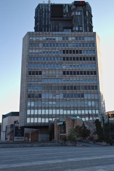 The photo shows a tall, rectangular modernist building in Ljubljana, Slovenia. Its facade is dominated by rows of square windows framed in concrete, giving it a rigid, grid-like appearance. Near the middle, two small flags hang inside one of the windows. At the base of the building, a mix of brick structures and greenery soften the stark geometry. Several national flags stand on poles to the right. The pavement in the foreground is patterned with repeating arcs, leading the eye toward the structure. At the top, a darker section of metal cladding rises above the concrete with a red digital clock.