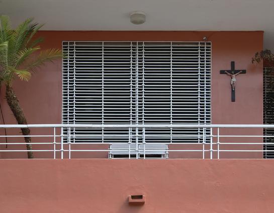 das foto zeigt einen abschnitt einer rötlich-erdfarbenen außenwand eines gebäudes. ein großes fenster mit einem weißen gitter ist mittig im bild. vor dem fenster befindet sich ein balkon mit einem ebenfalls weißen geländer. rechts neben dem fenster ist ein kruzifix an der wand angebracht. links im bild ist ein teil einer palme zu sehen. unten an der wand ist ein kleiner ausguss zu erkennen. über dem fenster oben an der decke ist ebenfalls mittig eine deckenlampe an der weißen decke zu sehen.
das gesamte bild wirkt ruhig und geometrisch.