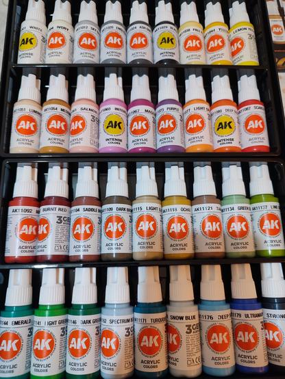 Bandeja de botellas de pintura acrílica de la marca AK Interactive organizadas en cuatro filas. Cada fila contiene varios frascos de colores diferentes, etiquetados con nombres como 'White', 'Ivory', 'Sky Blue', 'Lemon Yellow', 'Burnt Red', 'Dark Green', 'Turquoise', 'Ultramarine', entre otros. Las botellas son de plástico blanco con etiquetas en colores que reflejan el tono de la pintura.