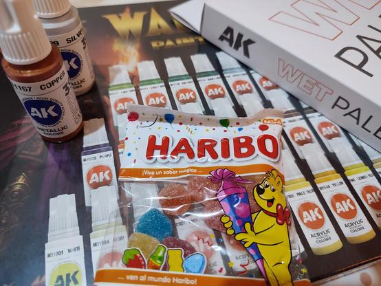 Variados productos de pintura acrílica de la marca AK Interactive, incluyendo frascos de colores metálicos 'Copper' y 'Silver', una caja de paleta húmeda (Wet Palette) y una lámina con ilustraciones de pinturas. En el centro de la imagen hay una bolsa de gomitas de la marca Haribo, con el oso amarillo característico sosteniendo un cucurucho de dulces. El fondo incluye imágenes de frascos de pintura organizados en filas.