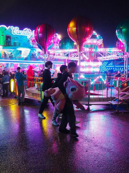 Picture of a group of teenage boys at a night funfair. Teen in the middle of the photo is carrying a big pink Stitch stuffed animal. Behind the teens there's a carousel and some other rides. Asphalt is wet so it reflects pink and blue lights from the rides.