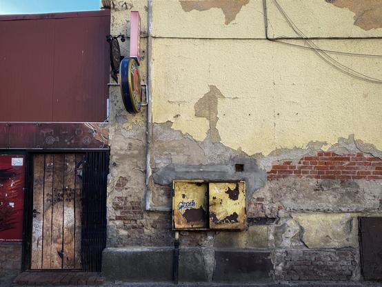 Eine verwitterte Gebäudefassade mit abblätternder gelber Farbe und freiliegenden Ziegeln. Eine Holztür mit einem schwarzen Gitter ist neben gelben und metallischen Gebrauchskästen zu sehen. Ein Schild mit Bierreklame hängt darüber, teilweise verdeckt. Das Gesamtbild ist schäbig und gealtert.