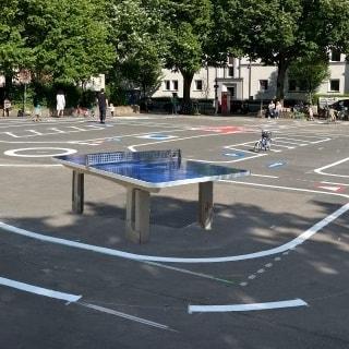 Das Bild zeigt eine öffentliche Spielanlage mit einem Tischtennisplatz im Vordergrund. Der Tischtennisplatz ist auf einem Asphaltboden mit weißen Markierungen eingezeichnet, die den Spielfeldrand und die Tischtenniszonen markieren. Der Tischtennisplatz ist mit einem blauen Tischtennisnetz ausgestattet. Im Hintergrund sind weitere Markierungen für andere Spiele wie Hopscotch zu sehen. Es gibt Bäume, die Schatten auf den Platz werfen, und im Hintergrund sind Gebäude und Menschen zu erkennen. Ein Fahrrad steht auf dem Platz, und einige Personen sind im Hintergrund zu sehen, die sich auf dem Platz oder in der Nähe aufhalten.