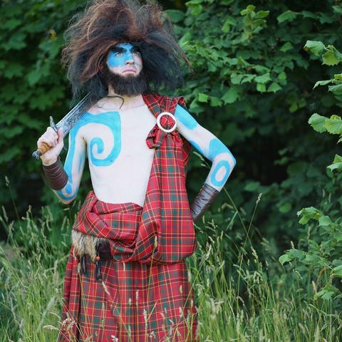 A man in a cosplay of Lord Macintosh from Brave, with red kilt and blue warpaint standing in a meadow
