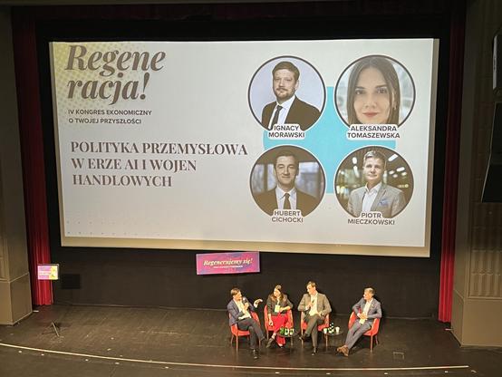 Widok na scenę głównej sali w Pałacu Kultury Zagłębia. Czwórka prelegentów siedzi w fotelach i prowadzi dyskusję, na ekranie za nimi wyświetla się nazwa kongresu, prelekcji (Polityka przemysłowa w erze AI i wojen handlowych) i ich zdjęcia
