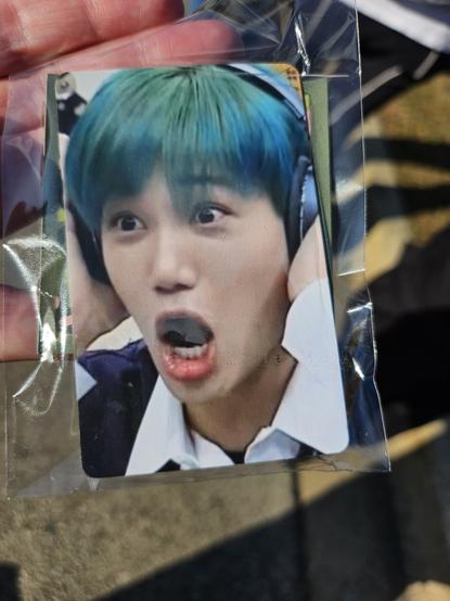 A hand holds a small plastic baggie with a wallet-sized photo card of EXO's Kai with teal hair, wearing headphones, mouth and eyes agape and shocked looking.