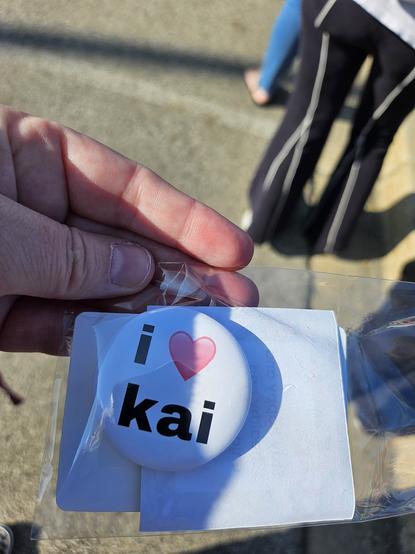 A hand holds a small plastic baggie with a homemade button that says "i 🩷 kai"