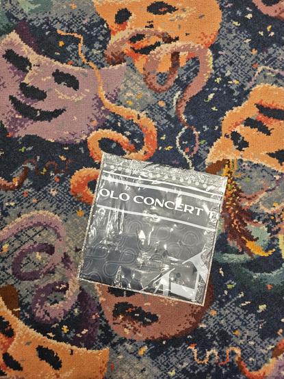 A folded KAION SOLO CONCERT bandana in its plastic packaging lays on the busy colorful carpet of a theater lobby.