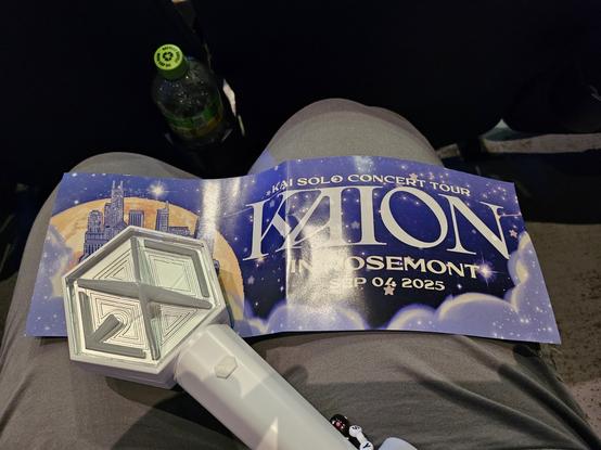 White EXO lightstick on top of a lavender and blue colored fan banner for KAION Rosemont on a person's grey-pantsed lap.