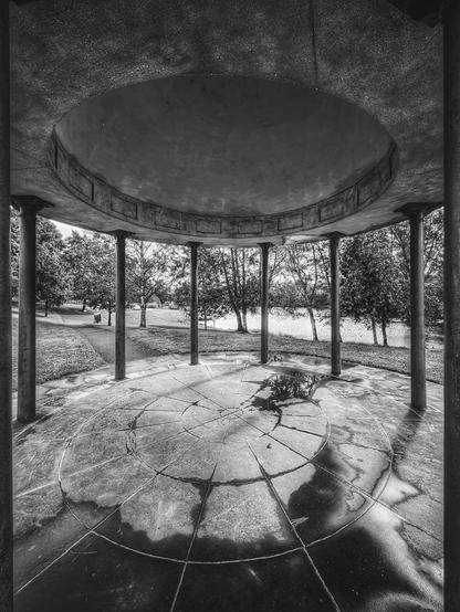 A monochrome, low-angle shot from inside a circular, open-air structure with a concrete floor and ceiling supported by several pillars. The floor has a circular, radiating pattern with a small puddle of water in the centre reflecting the sky and trees. Beyond the pillars, there's a grassy park with a path and a line of trees. The image has a calm, reflective, and slightly moody tone.