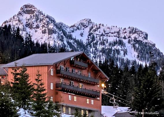 A winter evening scene at Alpental in Snoqualmie Pass, Washington. In the foreground stands a tall, rustic wooden ski lodge with balconies and glowing windows. A chairlift rises from the base, illuminated by a single yellow light. Behind the lodge, dramatic rocky cliffs rise steeply, covered in evergreen trees and deep snow, their jagged peaks outlined against a pale twilight sky. The composition contrasts the warmth of the man-made lodge with the rugged cold of the surrounding mountains.