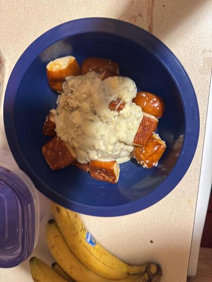 Blue bowl with pretzel bites covered in a creamy cheese sauce. The pretzel pieces are golden brown with a shiny surface, and the cheese sauce looks thick and slightly textured. The bowl is sitting on a countertop next to some ripe bananas and a plastic container with a purple lid. The overall setting looks casual and homey, likely in a kitchen.
