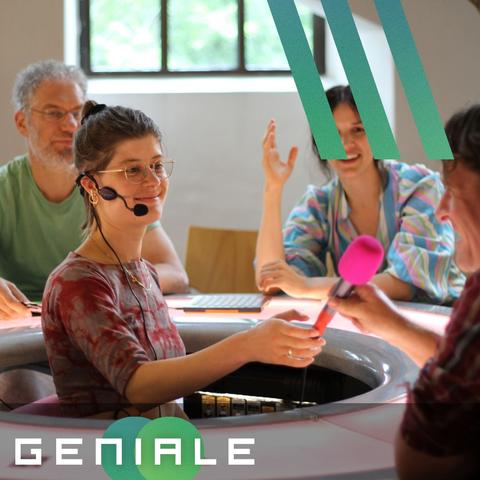 Foto aus einer Talkaoke-Runde: Eine Person sitzt in der Mitte eines ringförmigen Tisches und hält einer, der außen sitzenden, Personen ein Mikrofon hin.