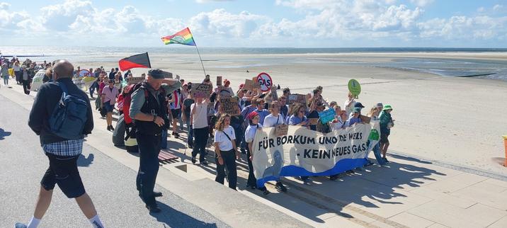 Das Bild zeigt eine Demonstration am Strand Borkums. Eine große Gruppe von Menschen marschiert mit Schildern und einem Transparent. Auf dem Transparent steht: "Für Borkum und die Welt - Kein neues Gas". Die Teilnehmer protestieren gegen die Errichtung neuer Gasinfrastruktur, wie aus den vielen Schildern mit entsprechenden Botschaften hervorgeht. Im Hintergrund ist das Meer zu sehen. Die Stimmung wirkt bestimmt und entschlossen.
