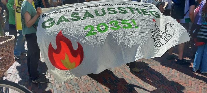 Das Bild zeigt ein Transparent der Demonstration. Auf dem Transparent steht in grüner Schrift „Fracking, Ausbeutung und Klimahölle – Gasausstieg 2035!“. Daneben ist eine stilisierte Flamme und eine Bohrinsel abgebildet. Das Transparent wird von mehreren Personen gehalten, deren Körper teilweise sichtbar sind. Der Hintergrund besteht aus einem gepflasterten Boden und weiteren Demonstranten.