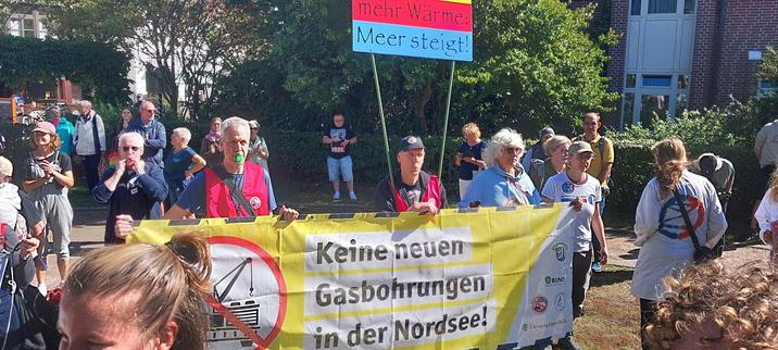 Das Bild zeigt eine Demonstration oder Kundgebung gegen neue Gasbohrungen in der Nordsee. Eine Gruppe von Menschen hält ein großes Banner mit der Aufschrift "Keine neuen Gasbohrungen in der Nordsee!" Die Teilnehmer scheinen besorgt über die Auswirkungen von Gasbohrungen auf die Umwelt und den Klimawandel zu sein.