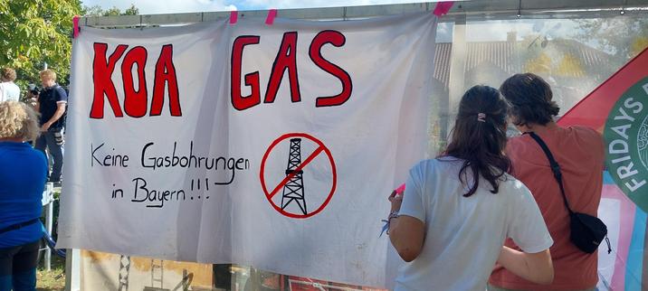 Das Bild zeigt ein Protestbanner mit der Aufschrift „KOA GAS – Keine Gasbohrungen in Bayern!!!“. Das Banner ist handgemalt und zeigt zusätzlich ein durchgestrichenes Bild einer Öl- oder Gasförderanlage. Zwei Personen, vermutlich Demonstranten, stehen vor dem Banner und betrachten es. Im Hintergrund sind weitere Menschen und ein Fotograph zu sehen.