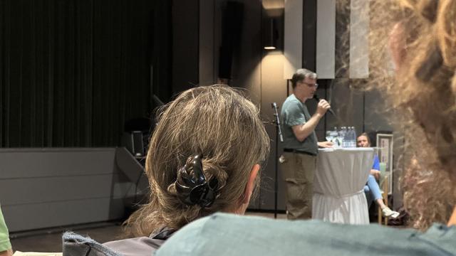 A speaker stands at a podium addressing an audience. In the foreground, the back of a person's head with a hair clip is visible. Several people are seated in the background, with a small table holding water bottles. The setting appears to be a conference